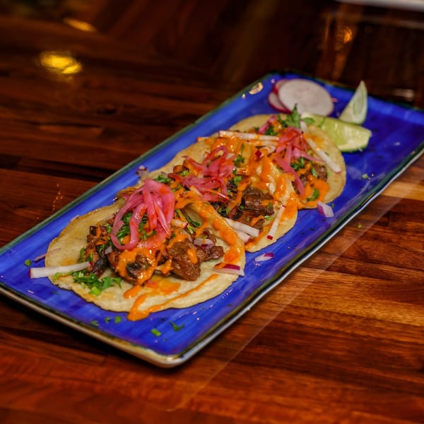 Three street tacos topped with meat, pickled onions, and orange sauce on a blue plate.