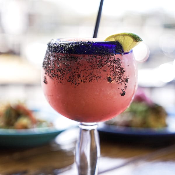 A vibrant pink margarita in a blue-rimmed glass with black salt and a lime.